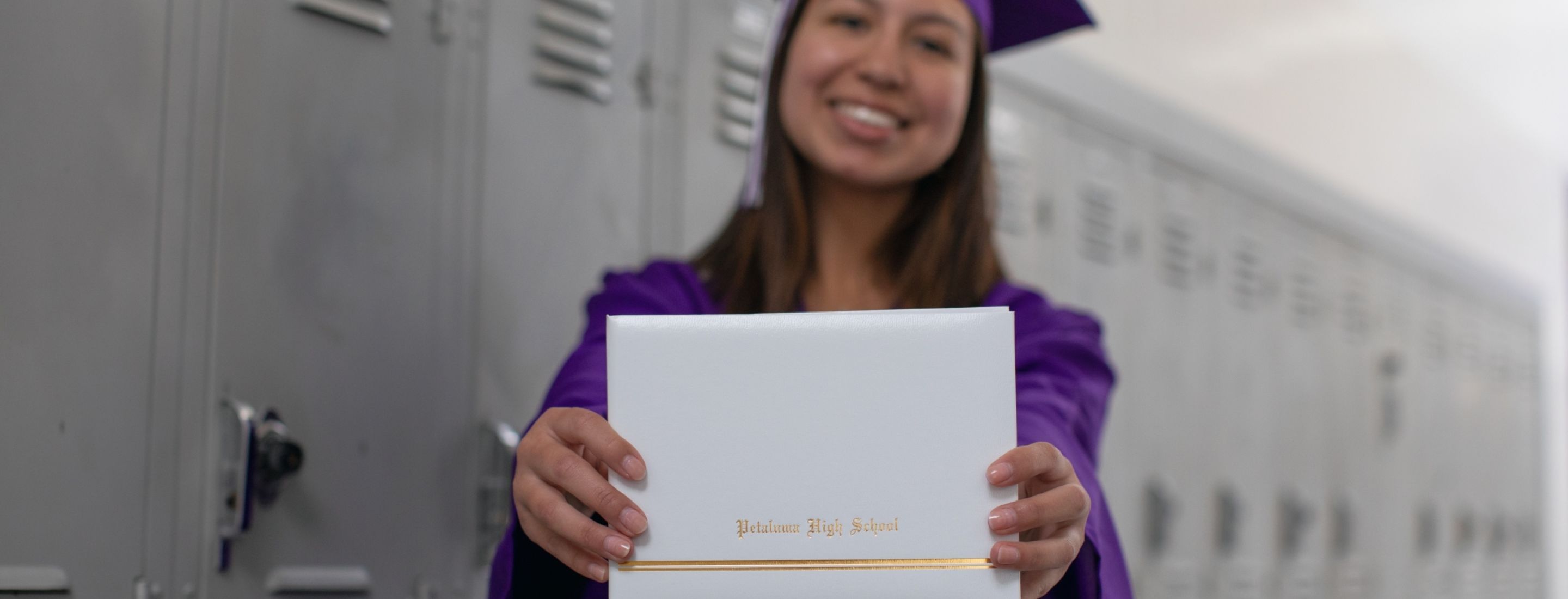 Petaluma High School student standing in front of gray lockers, wearing a purple graduation gown, and holding a diploma in front of the camera