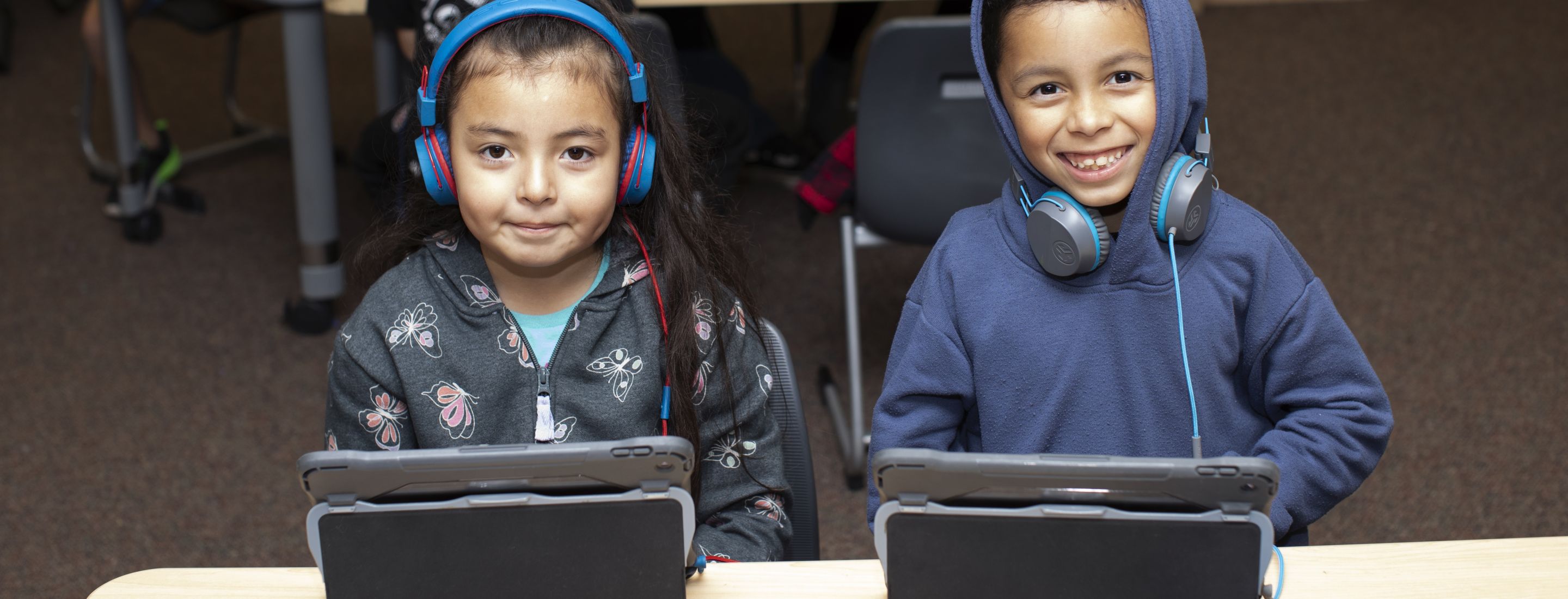 Two Penngrove students sitting at a desk, smiling for the camera. Both students are wearing headphones and using ipads.